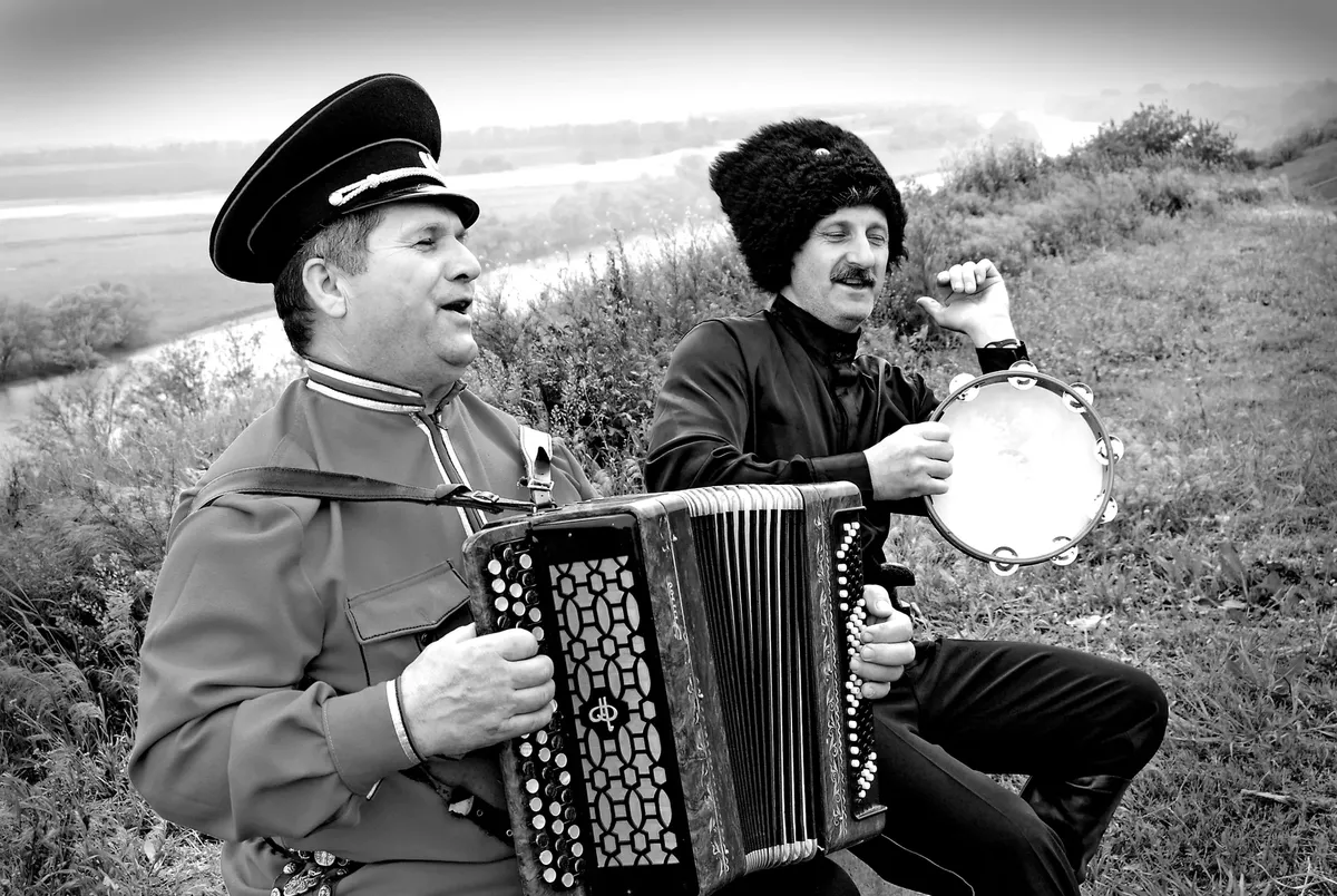 Black and white photo of men playing accordion and tambourine in traditional attire outdoors.